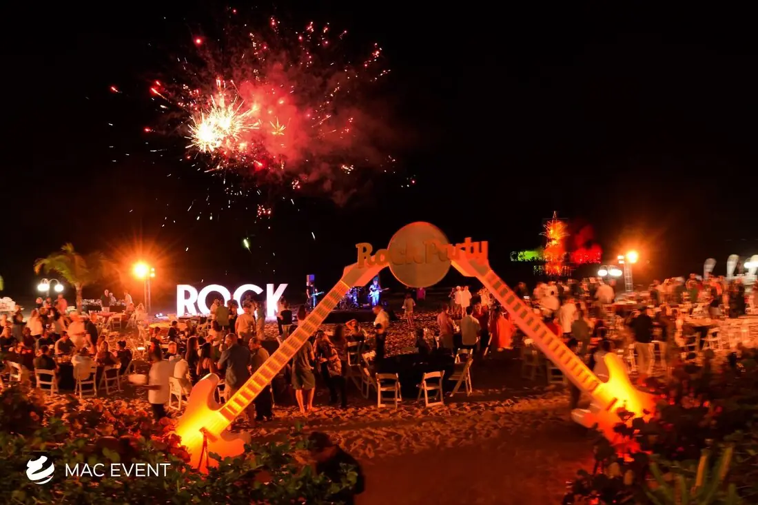 A lively beach party with a rock-and-roll theme, featuring a large guitar decoration, bright lights, and a crowd of guests enjoying fireworks overhead.