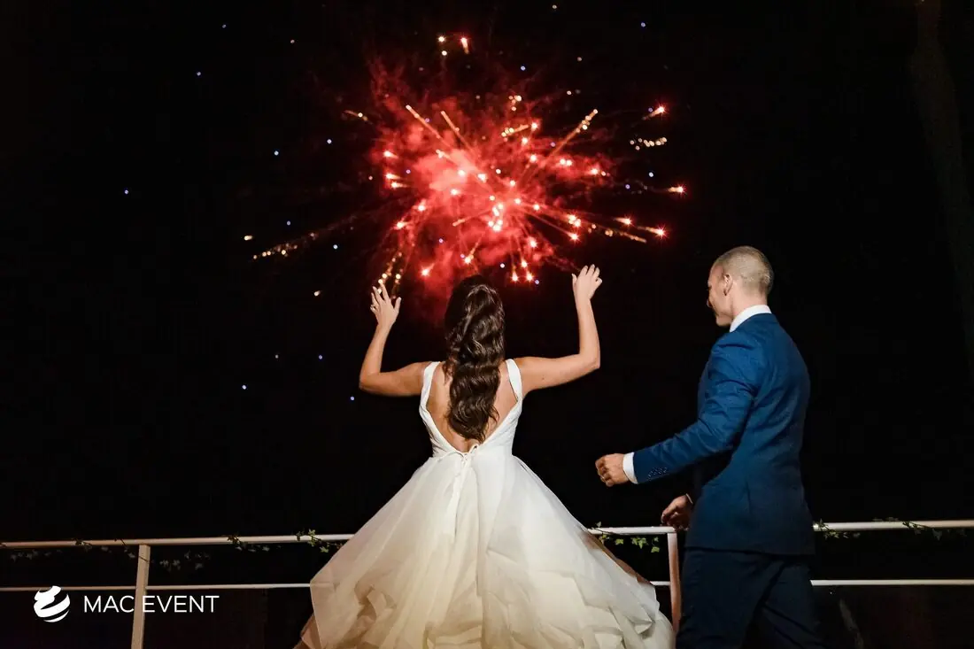 A bride in a flowing gown raises her hands in awe while watching a stunning red fireworks display with her groom at an evening wedding celebration.