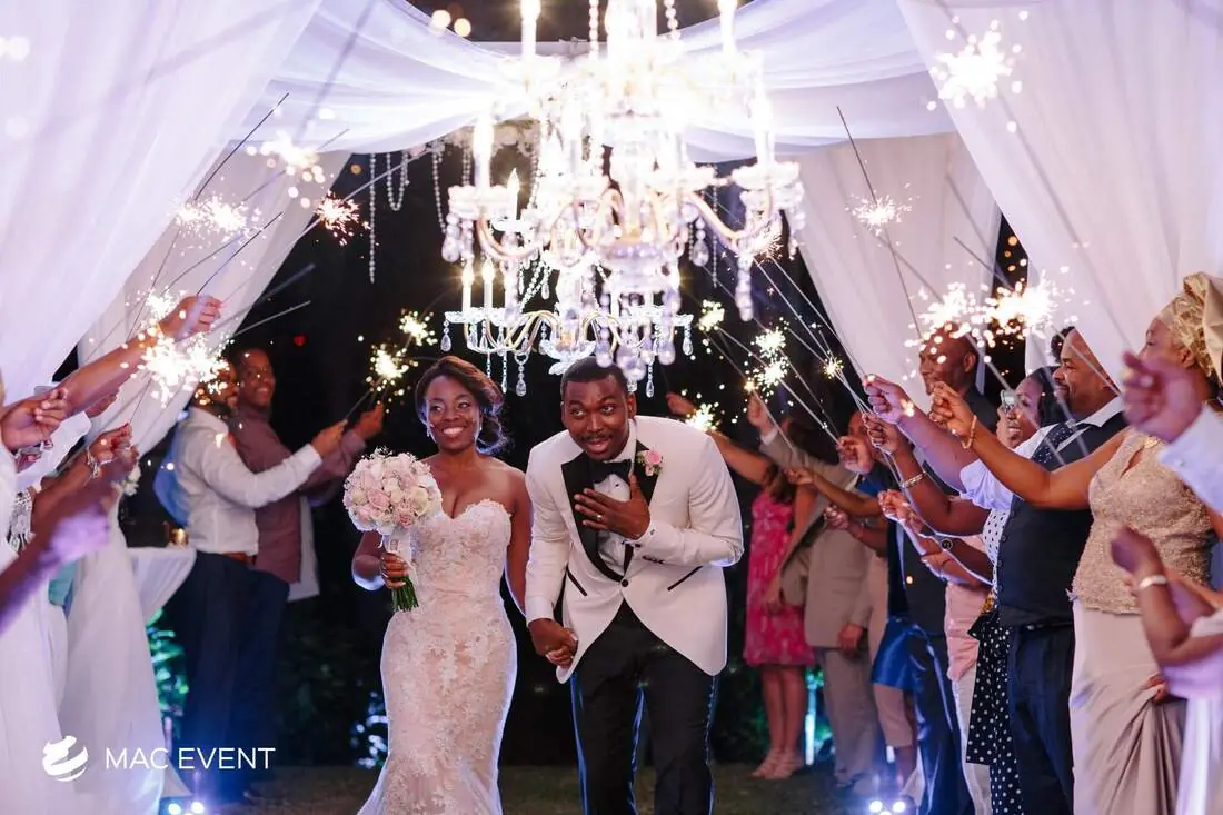 A bride and groom walk through a beautifully decorated entrance with chandeliers, smiling as guests wave sparklers around them in a grand wedding exit.