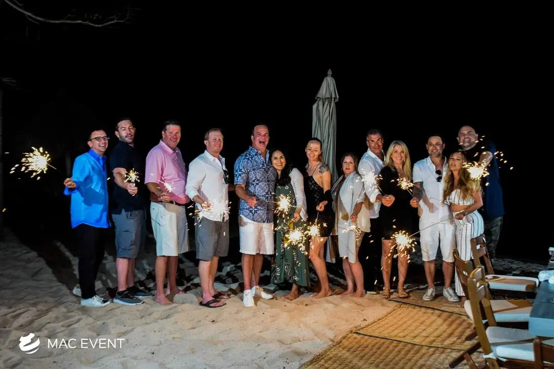 A group of wedding guests stands together on a sandy beach, holding lit sparklers and smiling at the camera, enjoying a festive nighttime celebration.